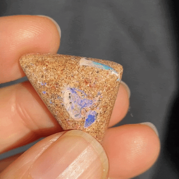 A close-up of a person's fingers holding a triangular piece of brown sandstone with streaks of blue and white opal visible within the stone.
