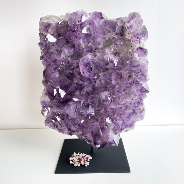 A large purple amethyst geode displayed on a black stand, with a cluster of smaller pink crystals at its base, against a plain white background.