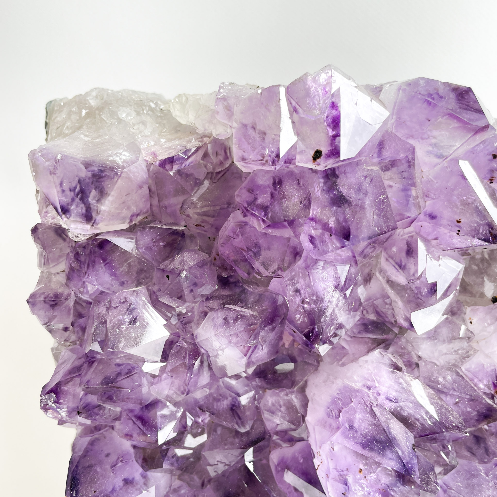 A close-up of a cluster of purple amethyst crystals with varying translucency and crystal sizes, set against a neutral background.
