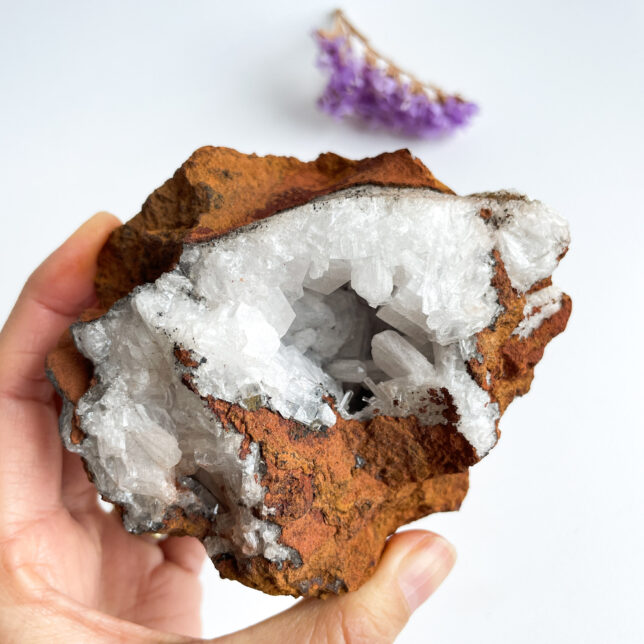 A person's hand holding a geode with quartz crystals inside, against a white background with a small purple amethyst cluster visible in the upper right corner.