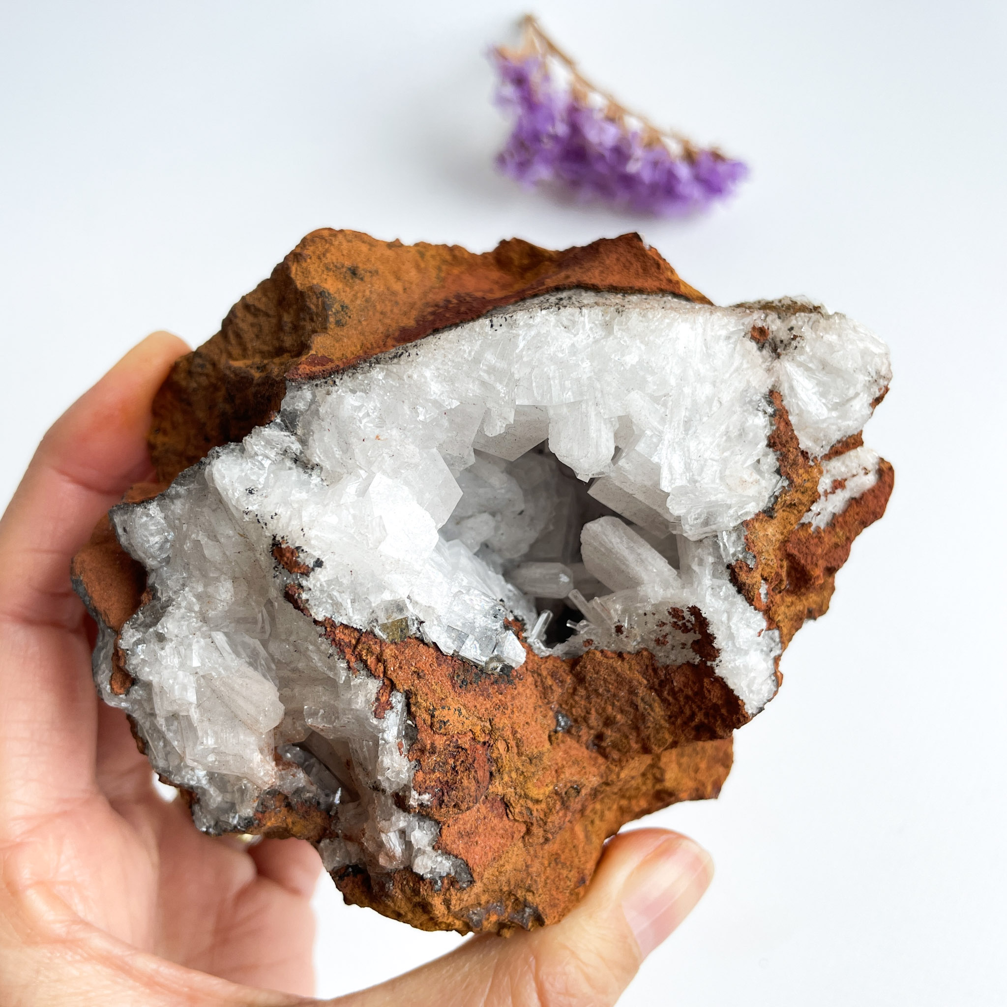 A person's hand holding a geode with quartz crystals inside, against a white background with a small purple amethyst cluster visible in the upper right corner.