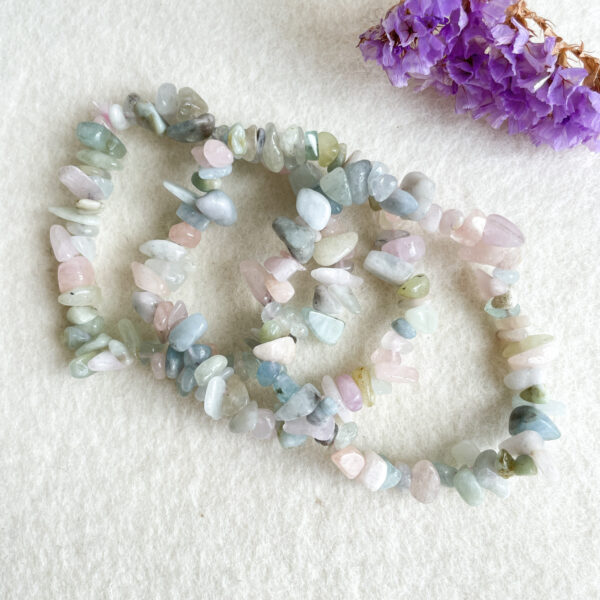 A strand of multicolored tumbled gemstone beads on a white surface, with purple dried flowers in the background.