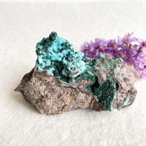 A piece of rough mineral rock with patches of bright turquoise crystals on its surface, placed next to a small bunch of purple flowers on a light background.