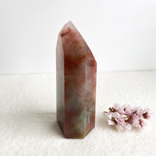 A polished, translucent crystal point with hues of red and green, standing on a white surface next to a small cluster of pink flowers.