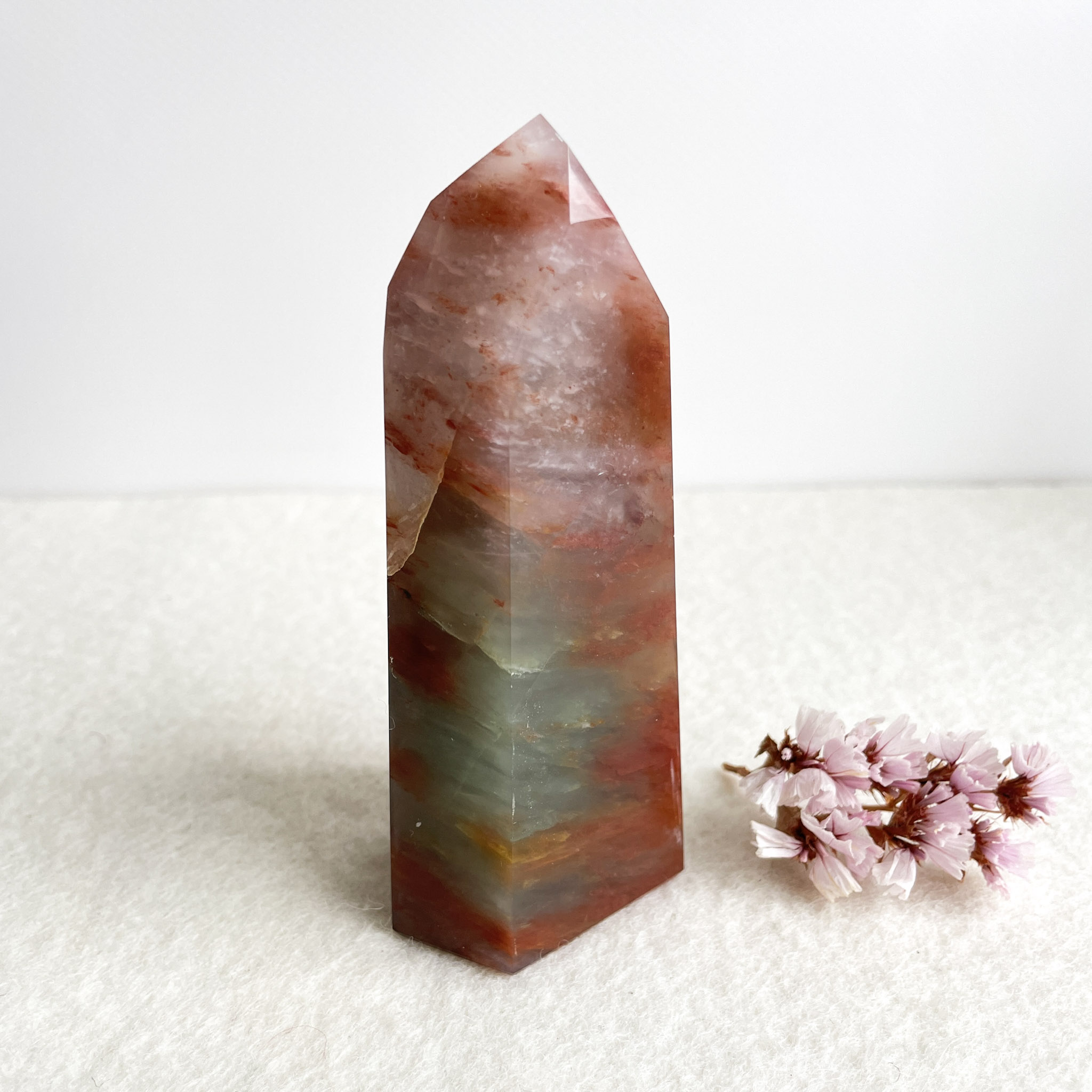 A polished multicolored crystal point standing on a white surface next to a small cluster of pink flowers, with a white background.
