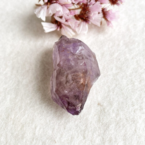 A close-up of an amethyst crystal on a textured white background with blurred pink flowers in the corner.