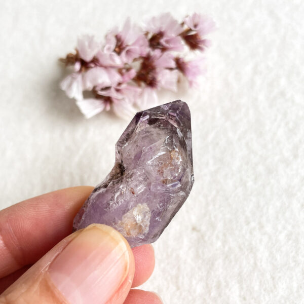 A close-up of a person's fingers holding a translucent purple amethyst crystal with a blurred background of soft pink blossoms on a white surface.