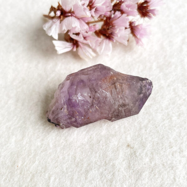 A raw amethyst crystal on a white textured background with a cluster of small pink flowers in the top left corner.