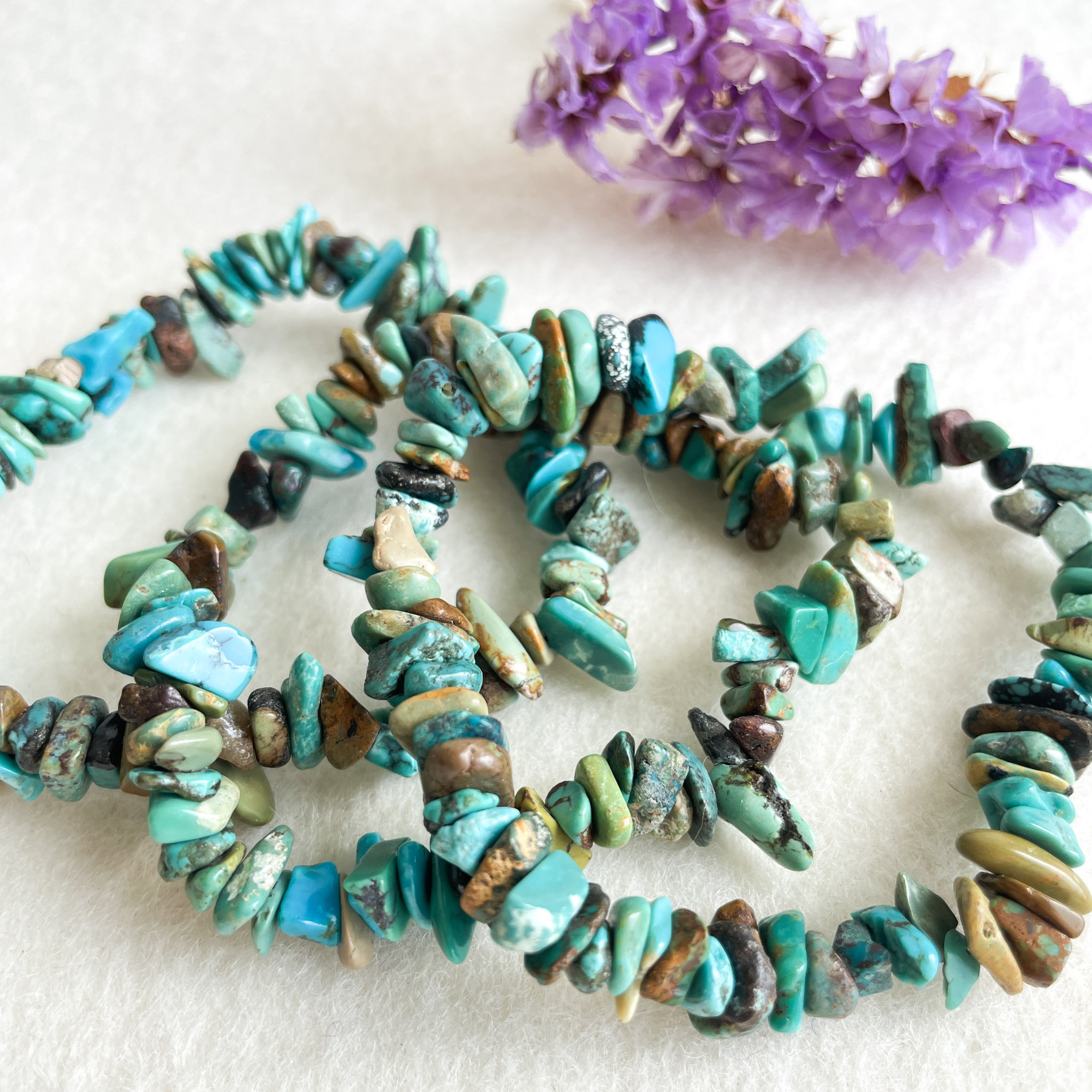 A necklace made of various shades of turquoise and brown gemstone beads arranged in a circle on a white background, with a sprig of purple flowers in the upper right corner.