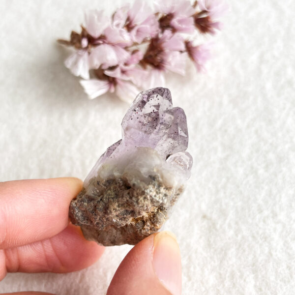 A hand holding a small, pointed amethyst crystal cluster with a white and beige background, and blurred pink cherry blossom flowers in the upper left corner.