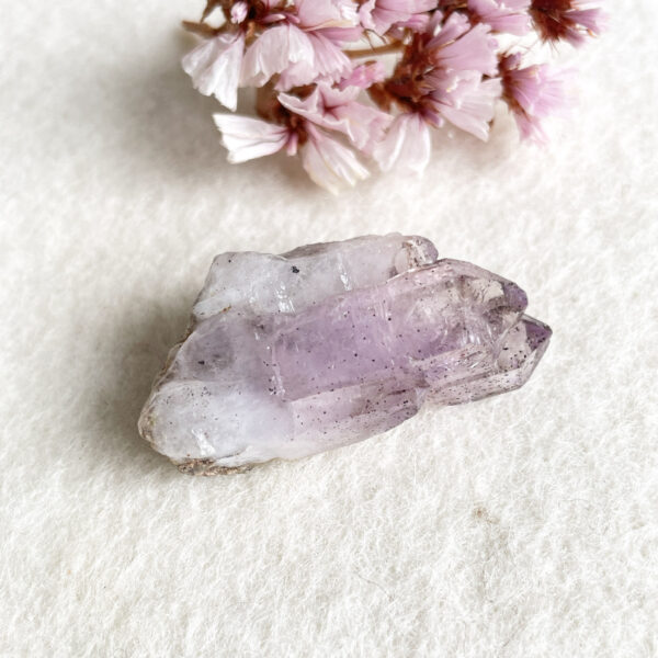A translucent purple amethyst crystal on a white textured background with a cluster of pink flowers in the top left corner.