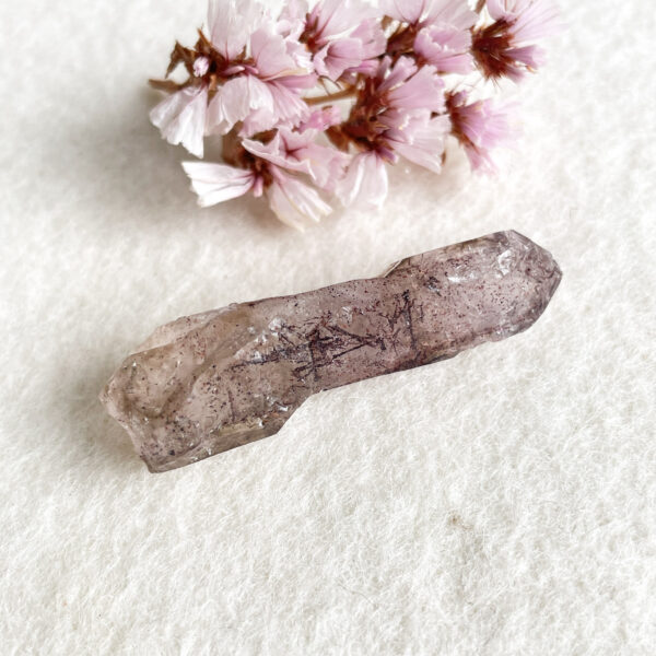 A translucent smoky quartz crystal lies on a textured white surface with a cluster of delicate pink flowers partially visible in the background.
