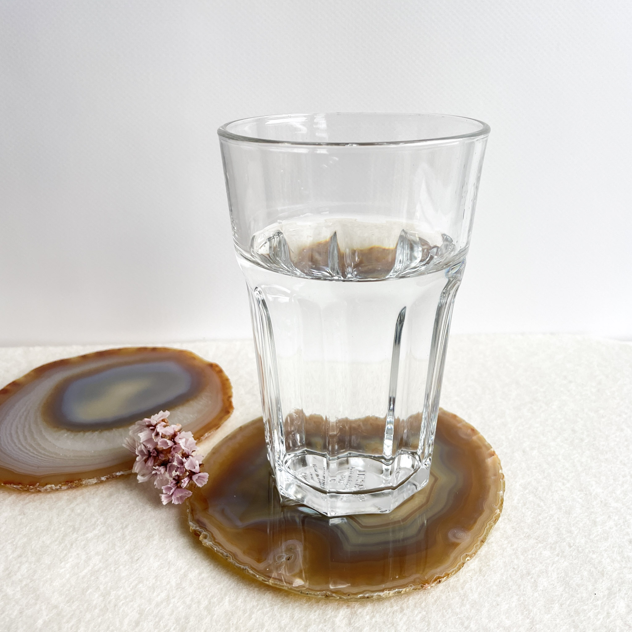 A half-full glass of water placed on a brown and white agate coaster, with a smaller slice of agate next to it adorned with small pink flowers, on a cream textured surface with a white background.