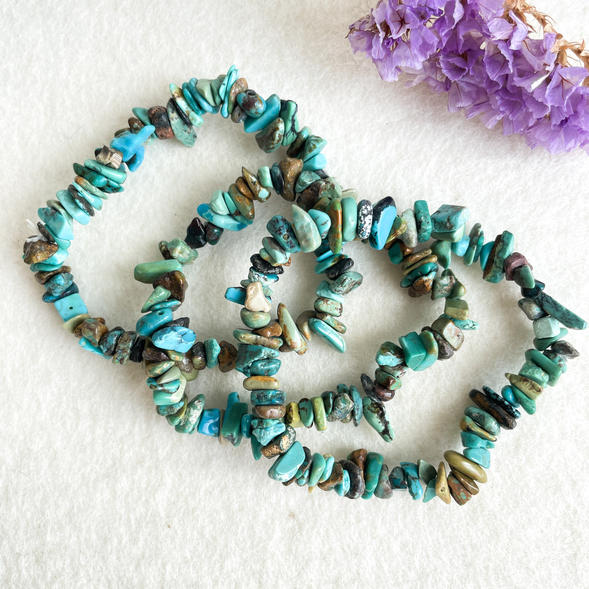 A string of turquoise beads in various shapes and sizes displayed in a spiral with a cluster of dried purple flowers in the top corner, all against a textured white background.