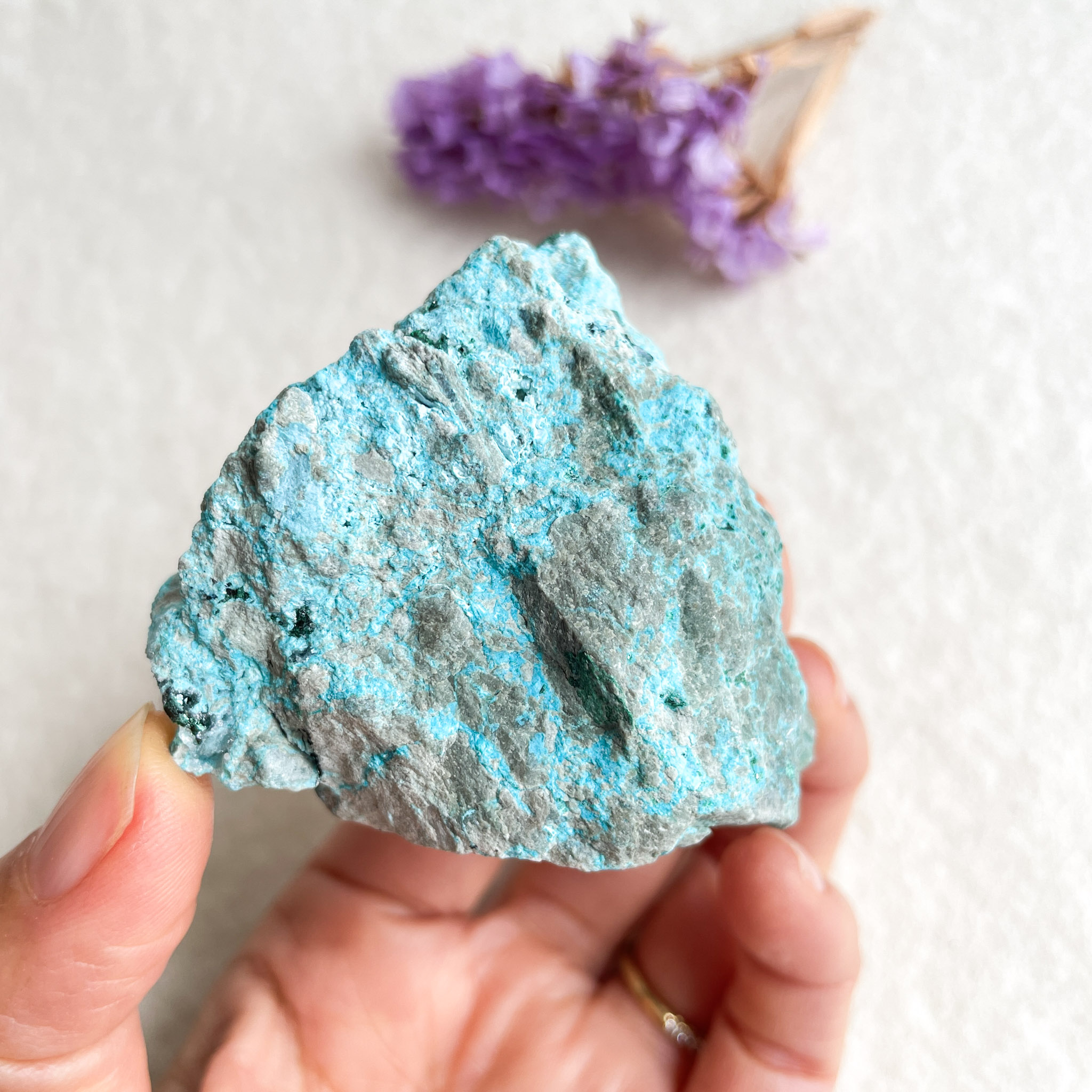 A person holding a blue mineral rock sample close to the camera, with a blurry background of white surface and a small bunch of purple flowers to the side.