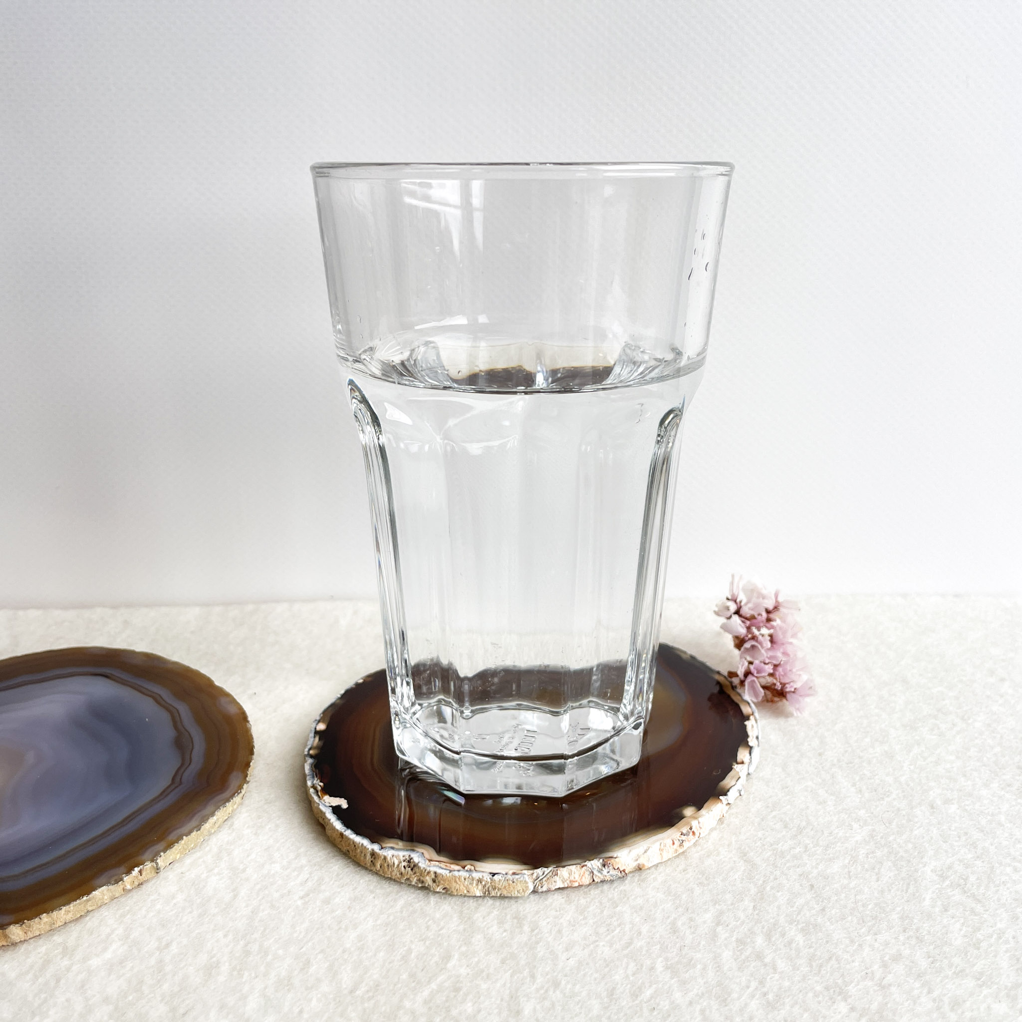 A half-full clear glass of water sitting on an agate coaster next to another similar coaster and a small pink flower on a textured white surface.