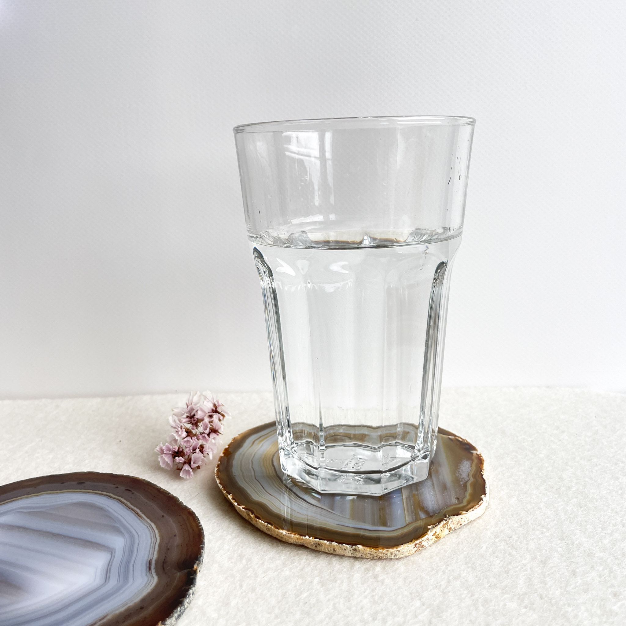A clear glass of water on a white surface with a small pink flower beside it, resting on a coaster made of a cross-section of an agate stone with gold edging.