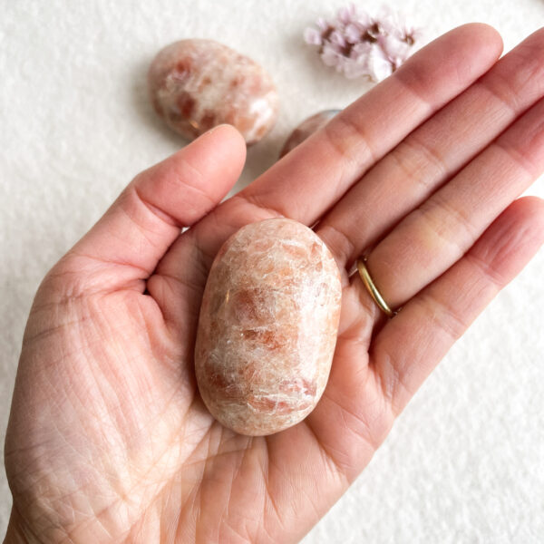 A person's open hand displaying a polished, oval-shaped pink stone, with another similar stone and some small pink flowers blurred in the background. The hand features a gold ring on the ring finger.