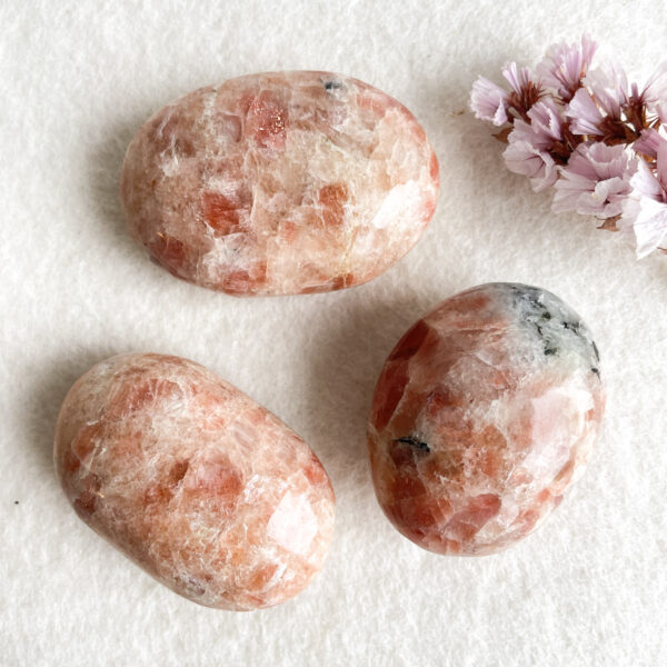 Three polished sunstone gemstones on a white textured background, accompanied by a small cluster of pale pink cherry blossoms in the upper right corner.