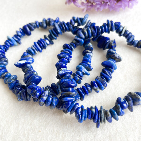 A string of lapis lazuli beads in various shapes and sizes arranged on a white surface with soft focus purple florals in the background.