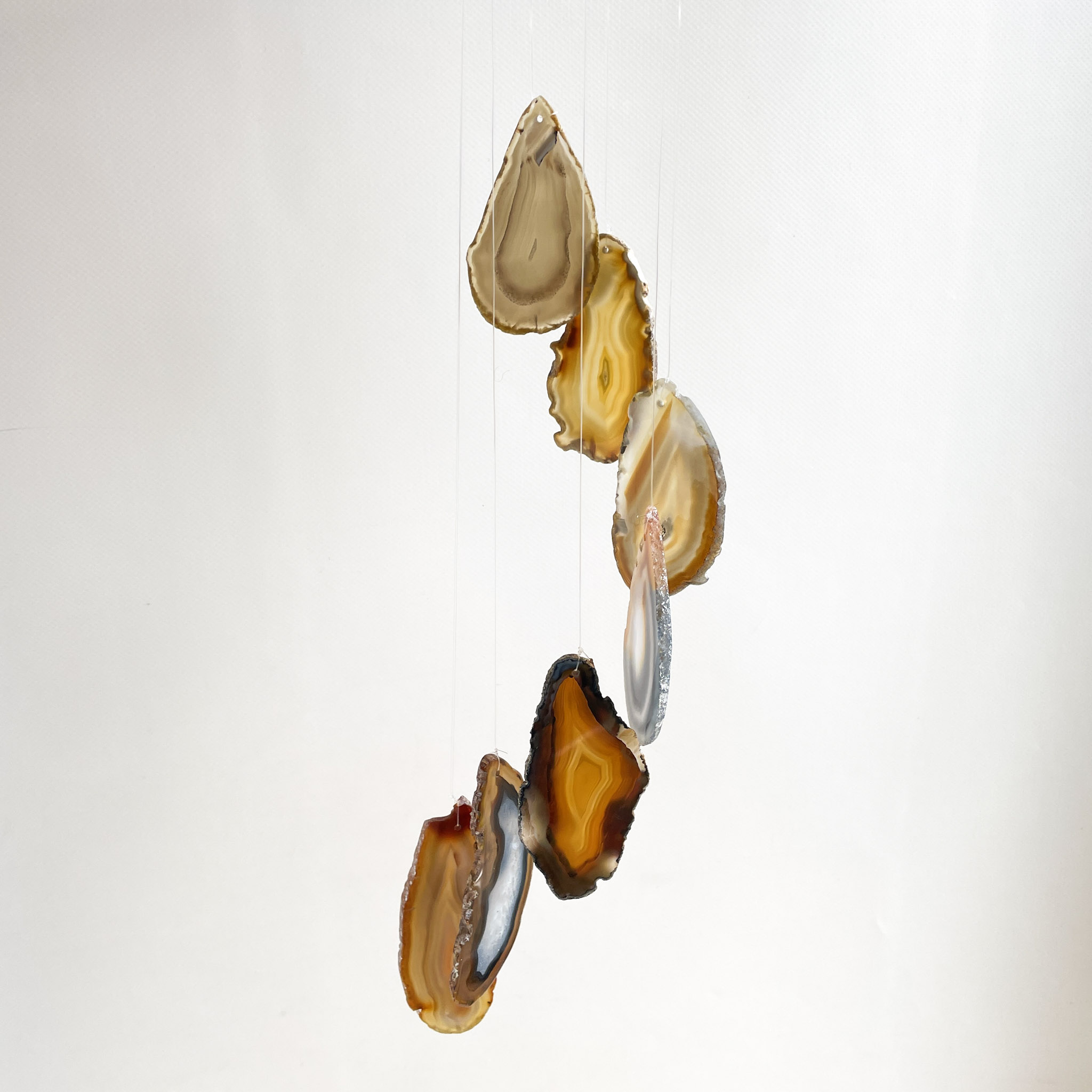 A group of sliced agate stones hanging by thin threads against a white background. The stones display layers of brown, beige, and creamy white with natural ridged edges.