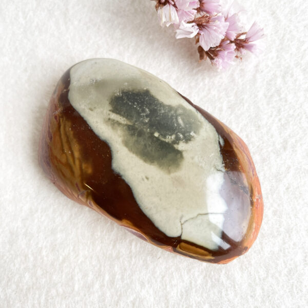 A polished multicolored gemstone with brown, white, and translucent layers, resting on a white textured surface next to small pink flowers.