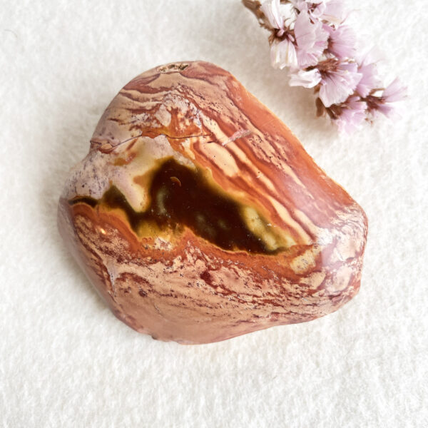 A polished multicolored stone with a glossy surface and varying shades of pink, cream, and brown, displayed on a white textured background next to a small cluster of pale pink flowers.
