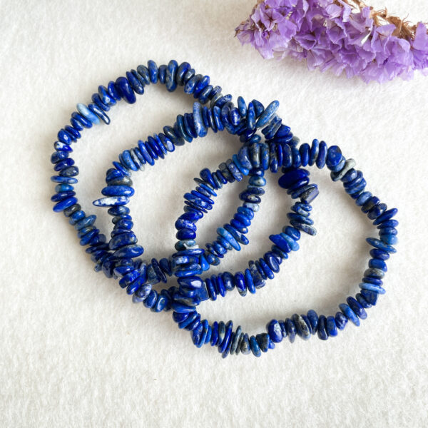 A spiral of lapis lazuli chip beads on a white surface with dried purple flowers in the background.
