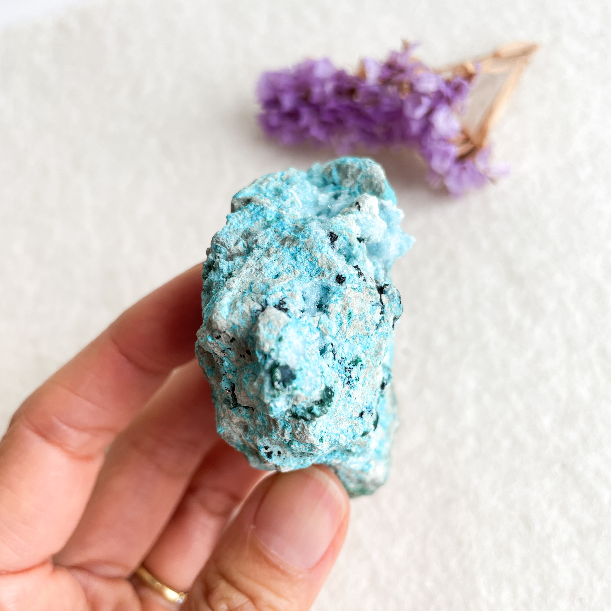 A person holds a bright blue turquoise mineral specimen with a blurred background of lavender flowers and textured white paper.