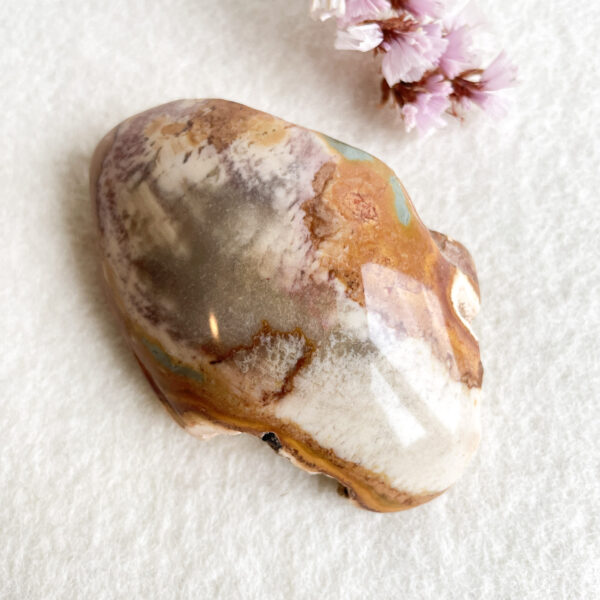 A polished multicolored stone with a mix of brown, white, and yellow hues resting on a white textured surface, with a few small pink flowers in the top right corner.