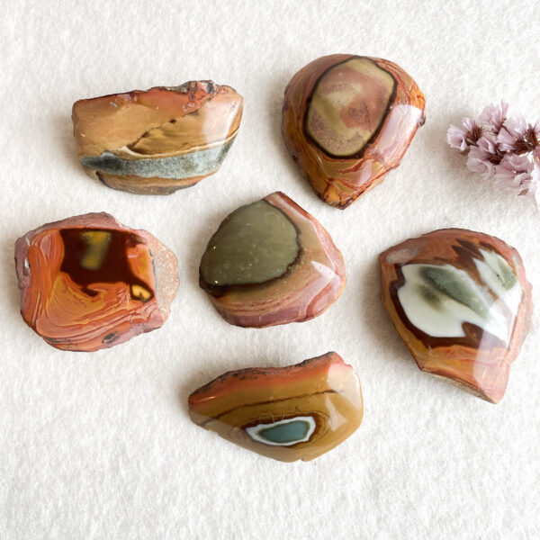 Seven polished stones with varying patterns of red, orange, beige, and green sit on a white textured surface, accompanied by small pink blossoms in the top right corner.