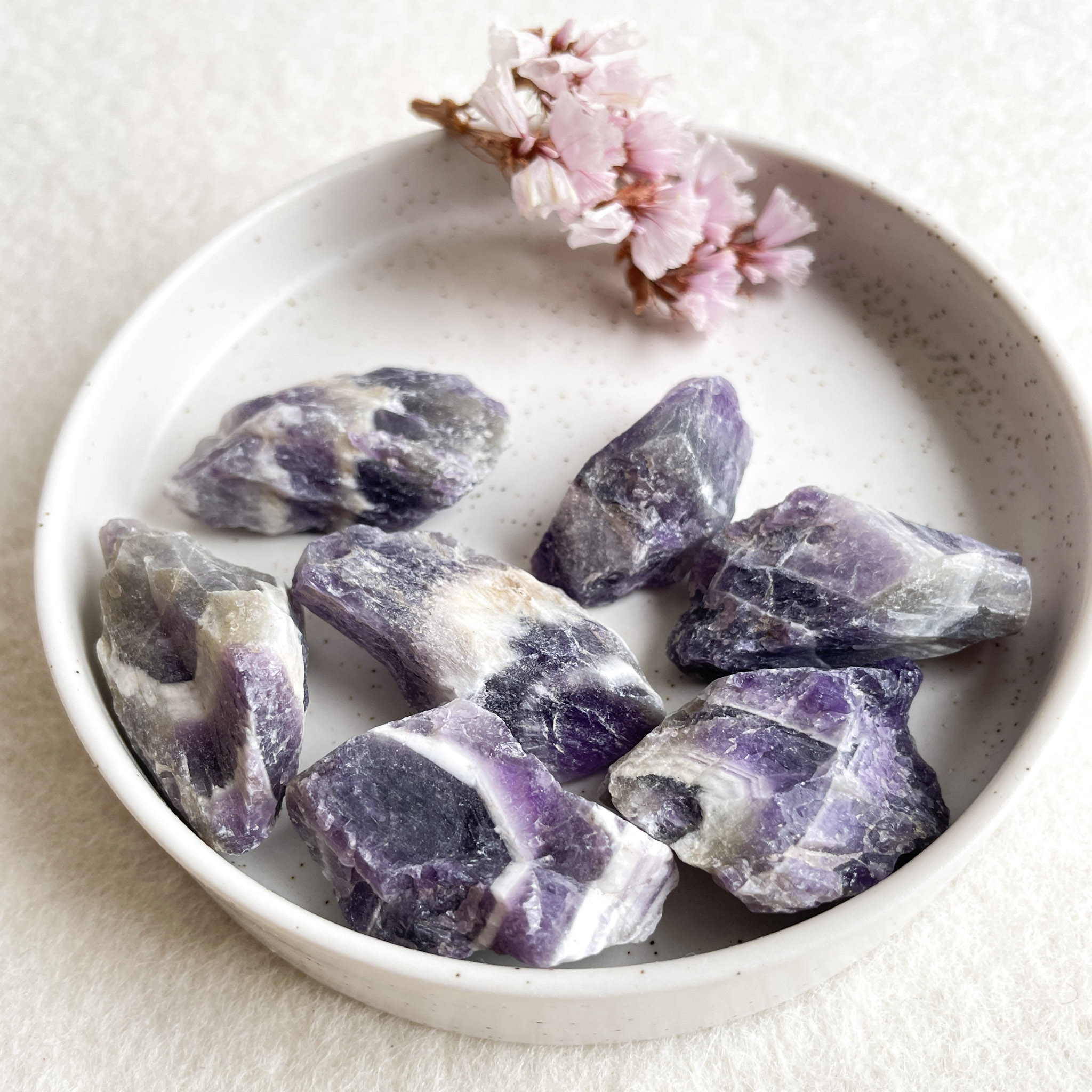 A white ceramic bowl containing rough amethyst crystals and a small cluster of pink flowers on a textured beige surface.