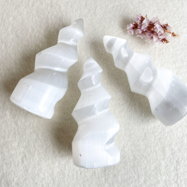 Three white, translucent selenite crystal towers of varying heights arranged in a diagonal line on a cream-colored surface, with a small cluster of pinkish dried flowers to their right.