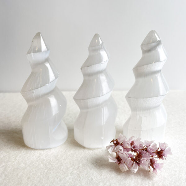 Three white, tiered selenite crystal towers in varying heights on a white background with a cluster of small pink flowers in the foreground.