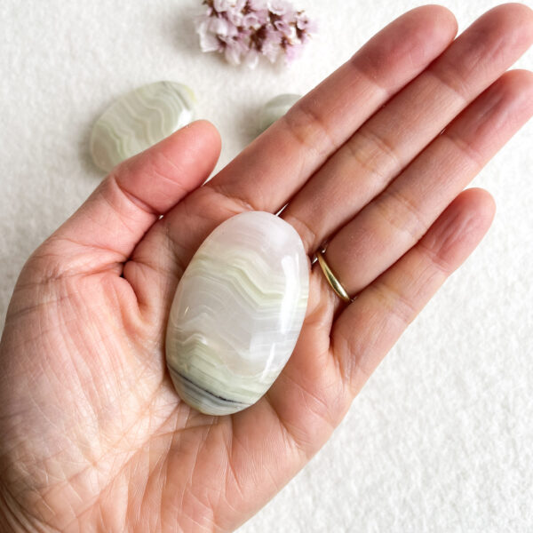 A person holding a polished, banded white agate stone in their palm, with two smaller stones and a cluster of pink flowers partially visible in the background on a white surface.