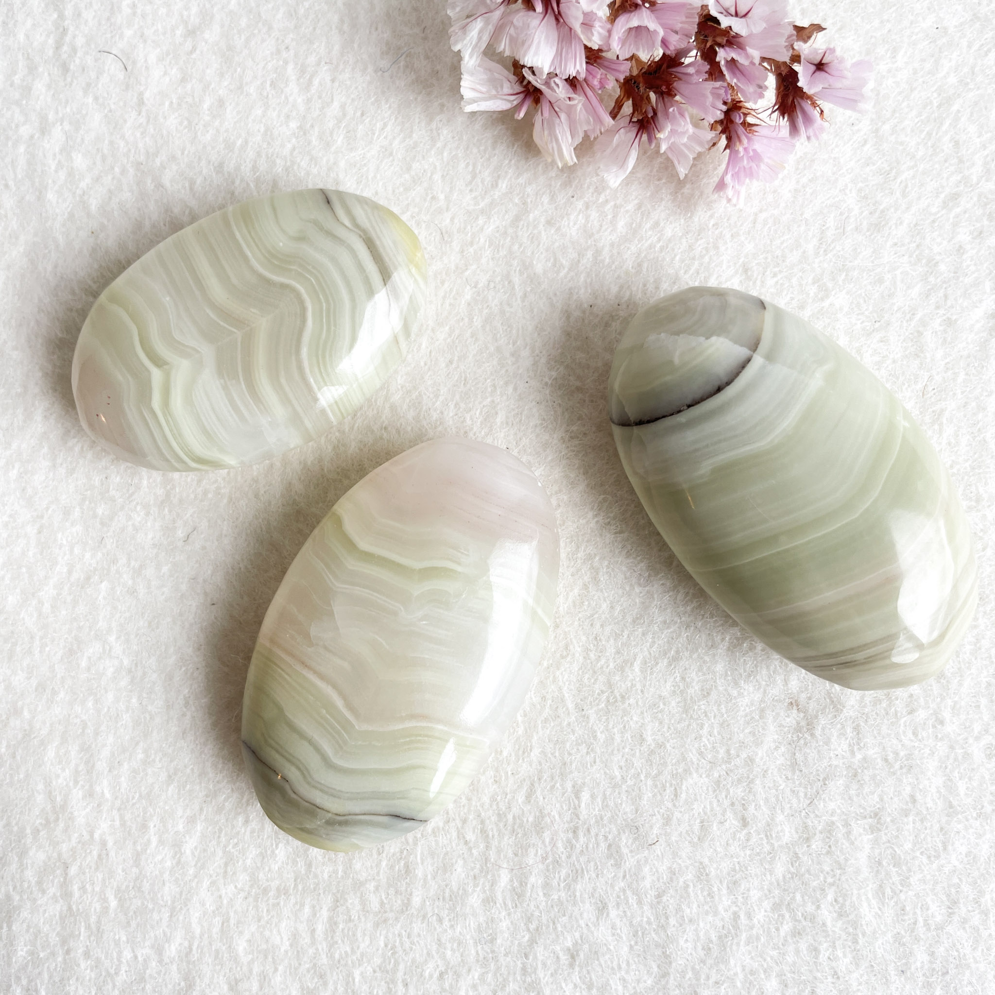 Three polished banded agate stones in shades of green and white on a white textured surface, with dried pink flowers in the background.