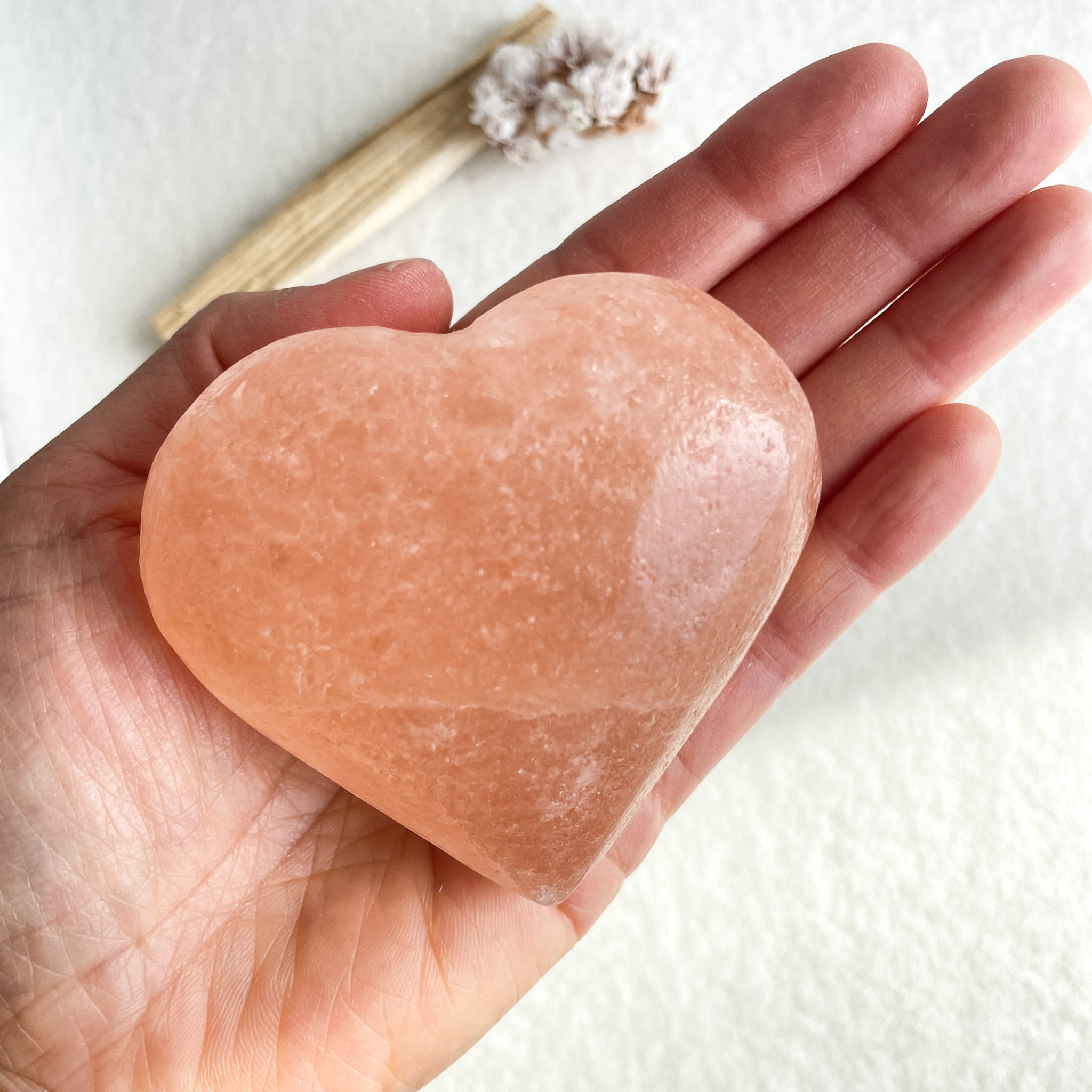 A person's hand holding a heart-shaped pink salt stone against a pale background with a dried flower and bamboo twig out of focus.