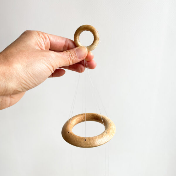 A hand holding a small wooden ring attached to strings suspending a larger wooden ring against a white background.