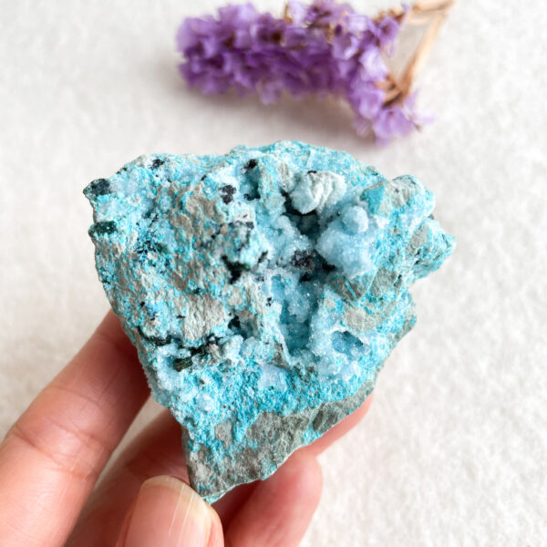 A person is holding a textured blue mineral specimen in front of a white background, with small purple flowers blurred in the background.