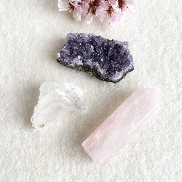 Three different crystal stones on a textured white background; an amethyst geode with deep purple crystals, a translucent clear quartz point, and a polished pink rose quartz obelisk, accompanied by pale pink dried flowers in the top right corner.