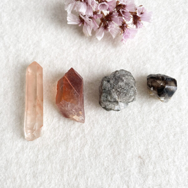 Alt text: Three crystals and a small cluster of flowers arranged on a textured white background. From left to right: a translucent pink crystal column, a reddish-brown pointed crystal, a rough gray stone, and a faceted black and white crystal, with a few delicate pink flowers placed at the top center.