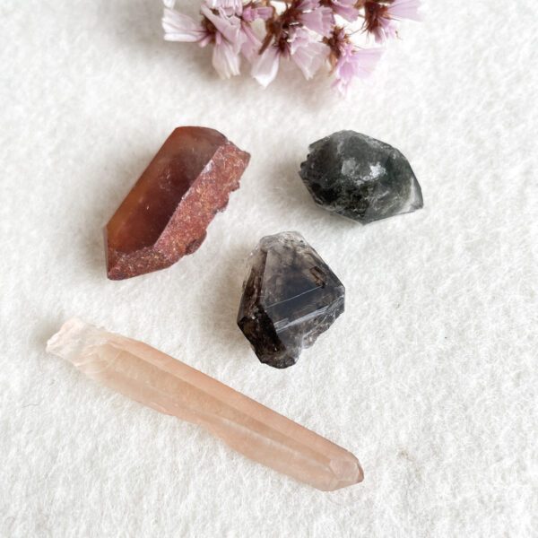 An assortment of rough gemstones and crystals of various colors and transparencies, along with a small cluster of pink flowers, arranged on a white textured surface.