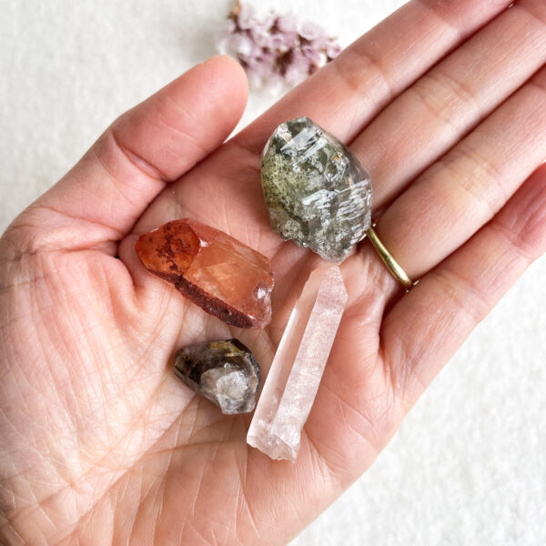 A hand holding a collection of various raw crystals and gemstones, with a blurred background of light-colored surfaces and flowers.