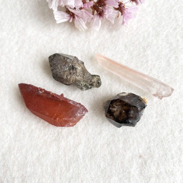An assortment of gemstones and crystal shards in varied colors displayed on a white textured background, accompanied by a cluster of light pink blossoms to the upper left corner.
