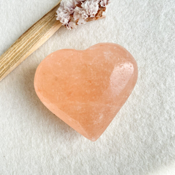 A heart-shaped pink Himalayan salt stone placed on a textured white surface next to a dried flower arrangement on a piece of driftwood.