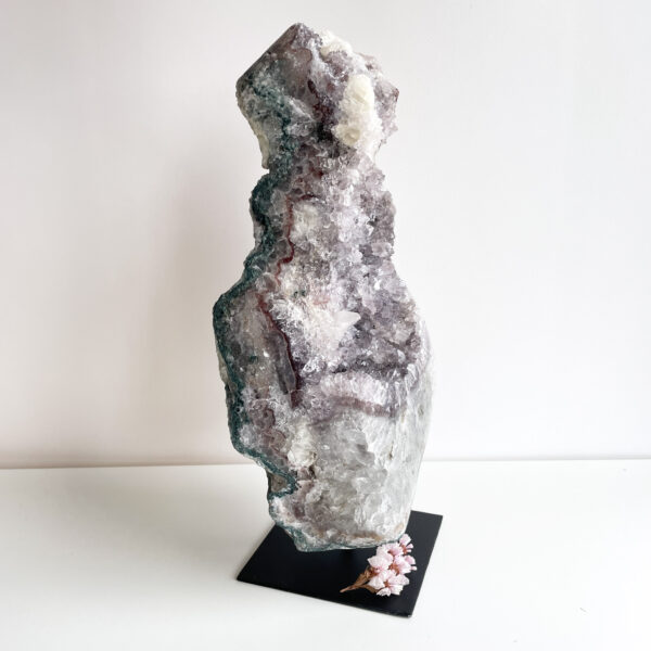 A large geode rock with crystalline structures on a black display stand with small pink flowers at the base, set against a plain white background.