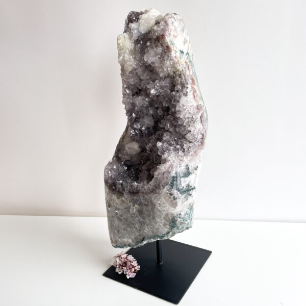 A large geode with a crystalline interior on a black display stand, with a small pink crystal flower on the base against a white background.