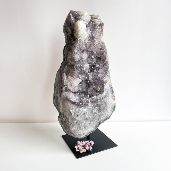 A large geode with sparkling purple and white crystals on a black display stand, with small pink blossoms placed at the base, set against a white background.