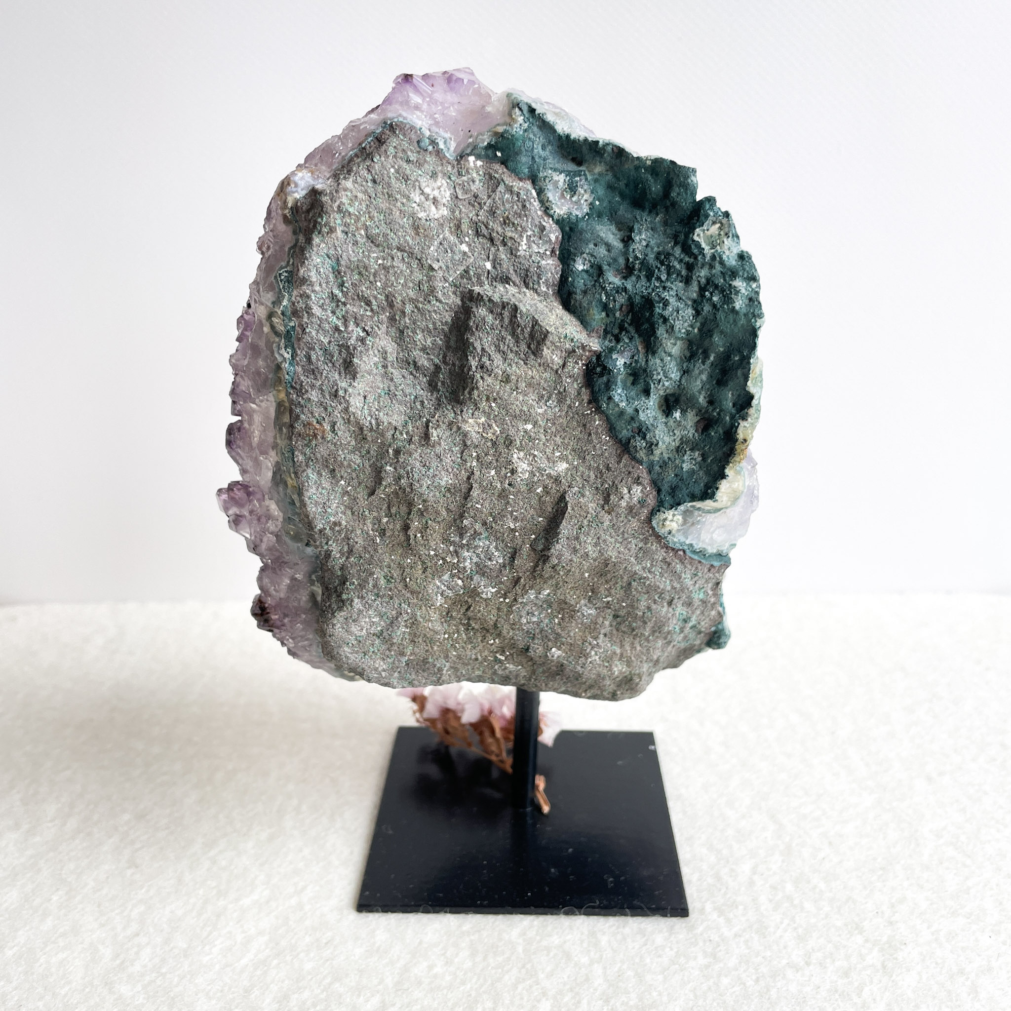 A large geode displayed on a black stand, featuring rough outer rock surface with an interior lining of crystalline amethyst and pockets of blue-green mineral deposits.