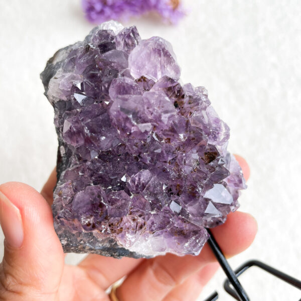 A hand holding a chunk of amethyst crystals with a blurred background of white surface and small purple flowers.